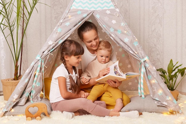 Indoor shot of mother with two little girls sitting together in wigwam mom with her children reading interesting book with fairy tale in peetee tent