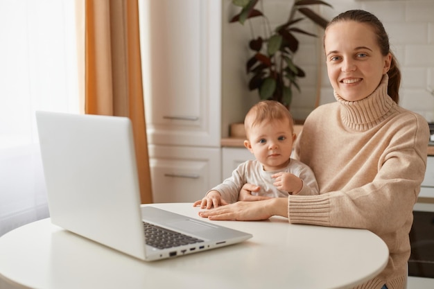 Indoor shot of optimistic delighted woman wearing casual style beige sweater sitting at table in kitchen with her baby daughter, working at laptop while looking after kid, looking at camera with smile