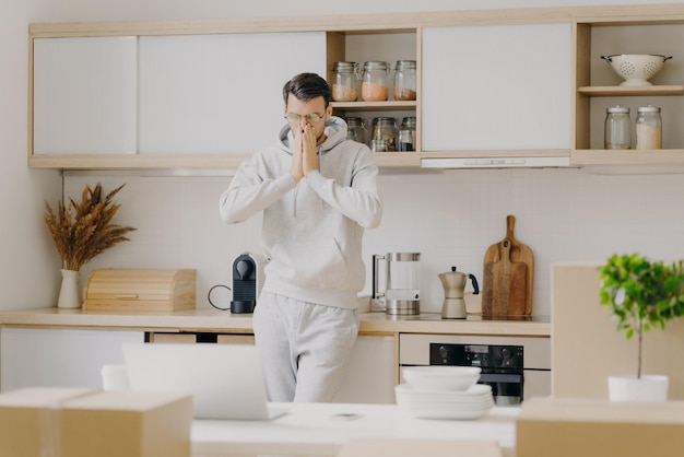 Indoor shot of pensive man works on family budget looks attentively at laptop computer poses in kitchen with unpacked boxes rents new house dressed in casual sweatshirt works remotely at home