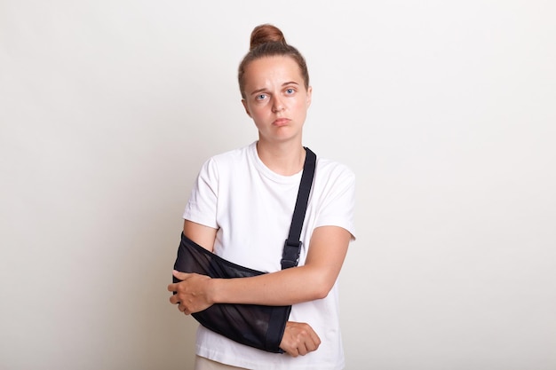 Indoor shot of sad sick woman with bun hairstyle wearing casual t shirt holding her painful arm in sling looking at camera with sadness and sorrow posing isolated over white background