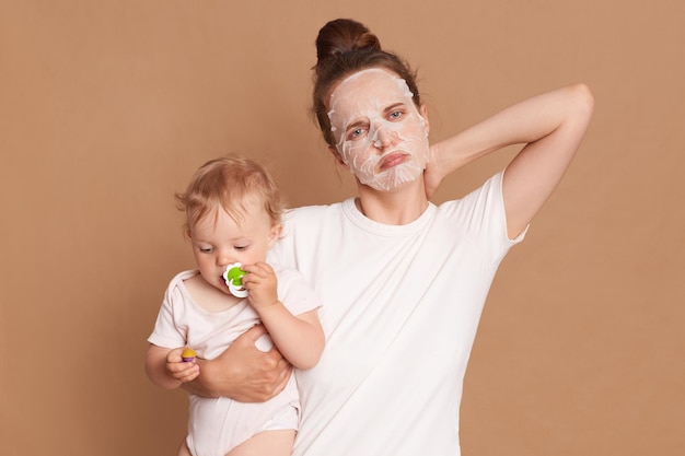 Indoor shot of sad unhealthy woman wearing white t shirt doing cosmetic procedures feels unwell has pain neck looking at camera with sad expression holding baby daughter in hands