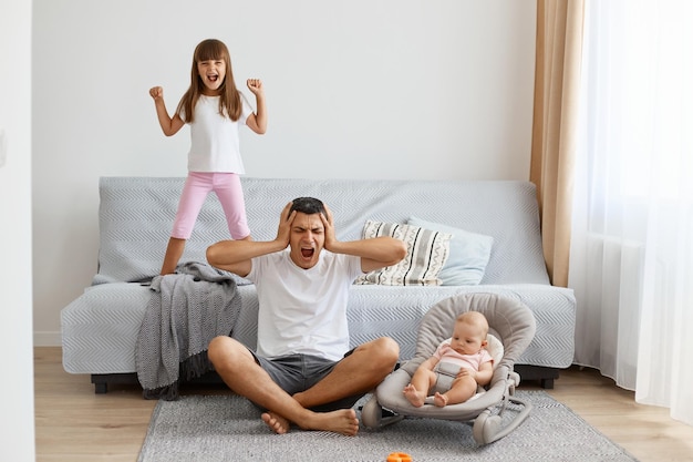 Indoor shot of sad upset depressed man wearing white casual style Tshirt and jeans short sitting on floor with his children covering ears being tired of screaming kids