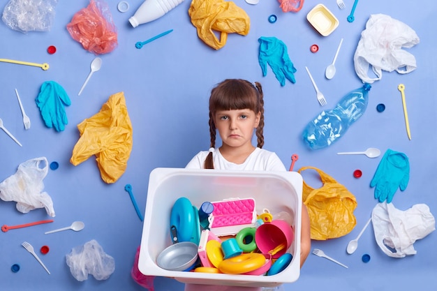 Indoor shot of sad upset little girl wearing white t shirt throwing away her plastic toys saving environment posing against blue wall and much plastic garbage around