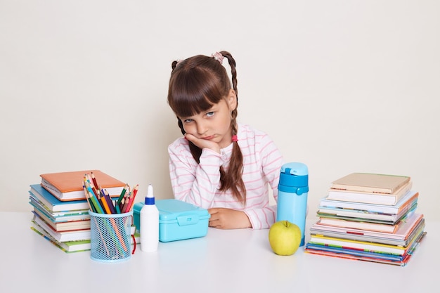 Indoor shot of sad upset little schoolgirl with dark hair and braids sitting at table surrounded with books and keeps hand under chin being bored and exhausted looking at camera