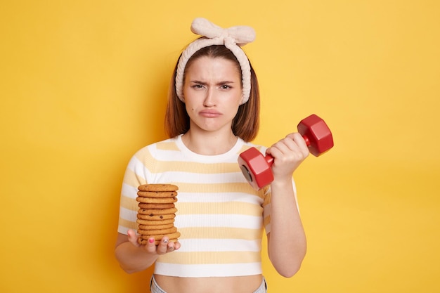 Indoor shot of sad upset woman with hair band holding in hands red dumbbell and cookies needs to burn calories looking at camera with unhappy expression posing isolated on yellow background