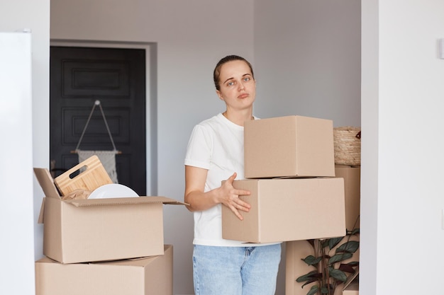 Indoor shot of sad woman wearing white casual style T-shirt and jeans standing with cardboard boxes with stuff, looking at camera, expressing sadness, being upset to leave her home.