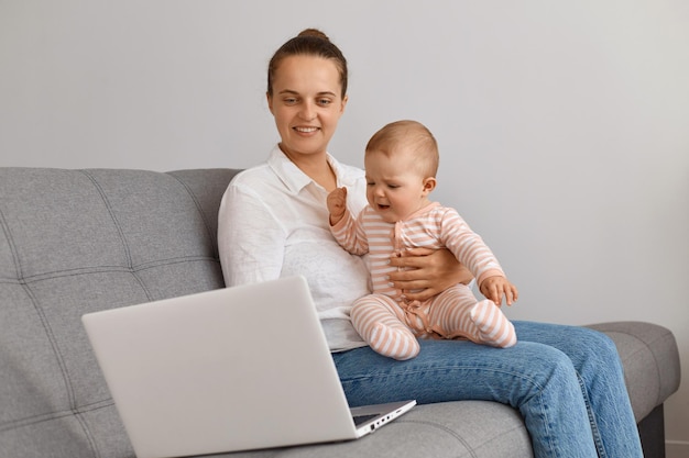 Indoor shot of satisfied female wearing white shirt and jeans sitting on sofa indoor and holding baby, looking at laptop screen, watching movie or having video call.