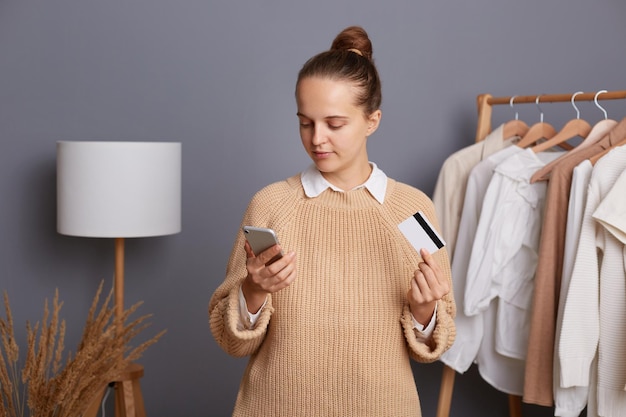 Indoor shot of serious concentrated woman holding cell phone and credit card and entering bank data while paying for her purchases standing in showroom near shelf with outfits