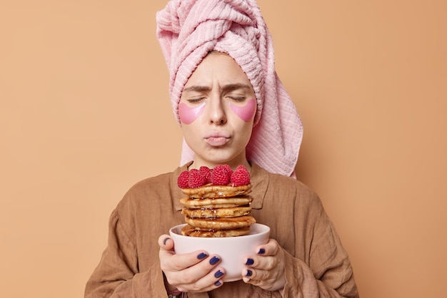 Indoor shot of serious young woman keeps eyes closed smells delicious pancakes with raspberries going to eat breakfast applies patches wears slumber suit and bath towel isolated over beige background