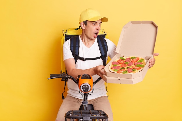 Indoor shot of shocked amazed courier man on bike opens carton pizza box and realize he is mixing up orders keeps mouth open wearing white t shirt and cap isolated over yellow background