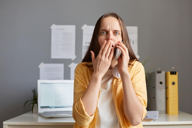 Indoor shot of shocked astonished Caucasian woman with brown hair sitting in office at her workplace and talking on mobile phone wearing yellow shirt hearing surprised news covering mouth with palm