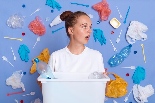 Indoor shot of shocked female holding box with plastic bottles has worried expression looks away at plastic garbage with widely opened mouth realizes seriousness of big environmental problem