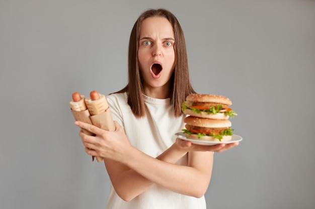 Indoor shot of shocked scared woman looking at camera with big eyes and open mouth keeps arms crossed holding sandwich and hot dogs keeps diet expressing astonishment