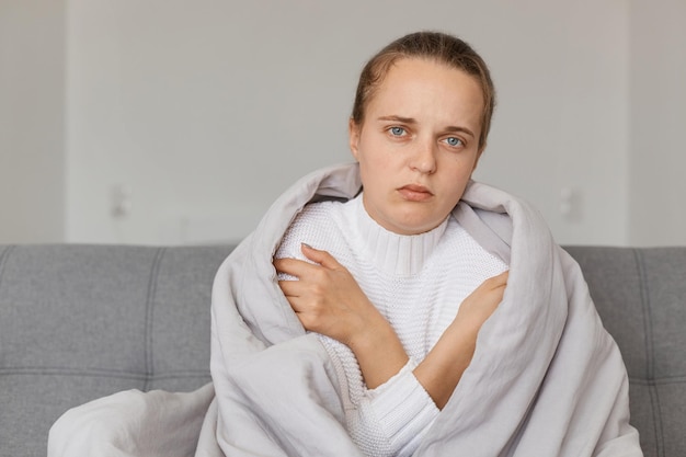 Indoor shot of sick unhappy woman being wrapped in plaid looking at camera with sad expression suffering influenza or grippe symptoms needs treatment