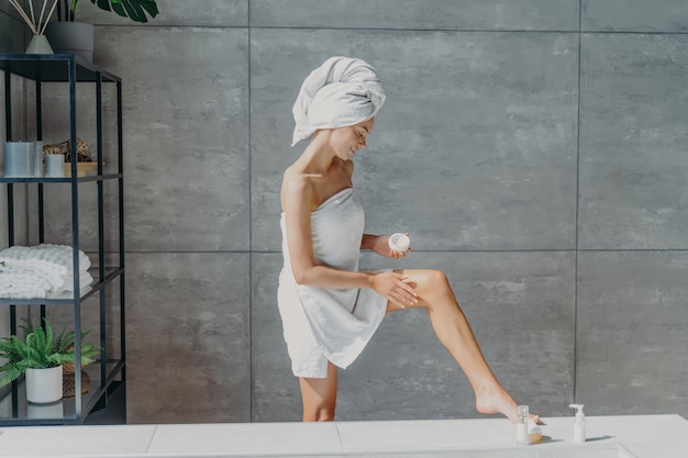 Indoor shot of slim female model applies leg cream stands wrapped in bath towel takes care of body and skin undergoes beauty treatments after taking shower poses in bathroom. Cosmetology concept