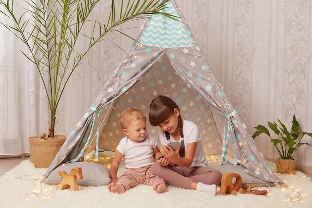 Indoor shot of smiling adorable little girls wearing white t shirts sitting on floor in peetee tent and playing with smart phone watching cartoons or playing games together