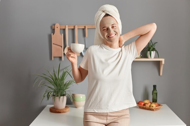 Indoor shot of smiling attractive positive woman with cosmetic mask on her face drinking coffee on the kitchen at home wakes up in good mood doing morning procedure for skin care