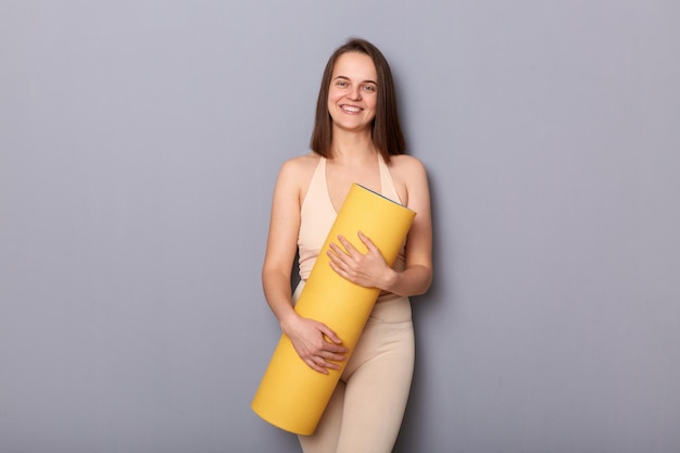 Indoor shot of smiling attractive woman with brown hair wearing beige top and leggins holding fitness mat isolated over gray background looking at camera enjoying her workout