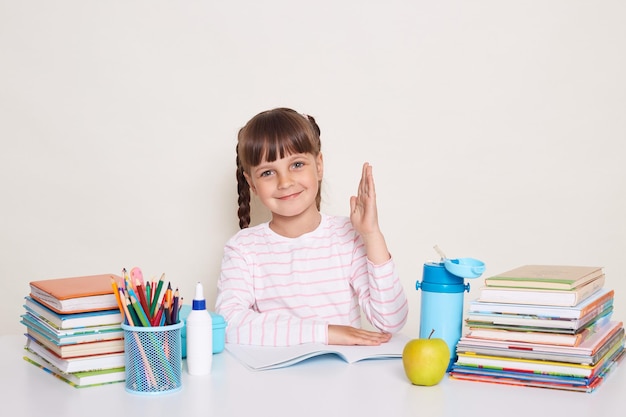 Indoor shot of smiling delighted little schoolgirl with dark hair and braids sitting at table surrounded with books and raised arm child want to answer during lesson