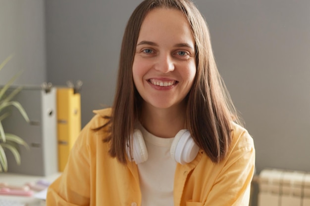 Indoor shot of smiling delighted young adult woman with brown hair wearing yellow shirt and headphones over neck sitting in office and looking at camera with happy expression