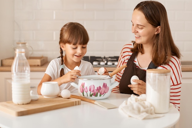 Indoor shot of smiling female with dark hair wearing brown apron baking homemade pastries with cute daughter in kitchen little girl adding eggs to dough family smiling enjoying cooking together