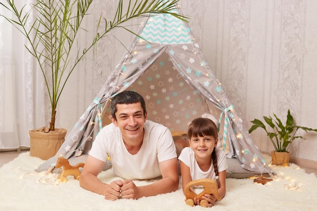 Indoor shot of smiling handsome Caucasian man wearing white t shirt lying on floor with his little daughter near her peetee tent looking at camera with positive expression