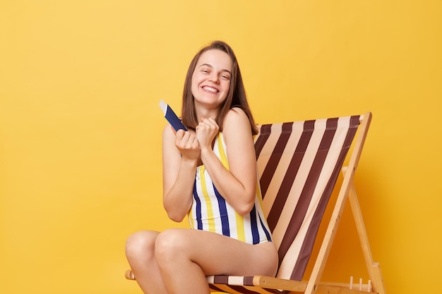 Indoor shot of smiling happy charming young woman wearing striped onepiece swimsuit sitting on wooden chair isolated on yellow background holding her passport enjoying longawaited vacation