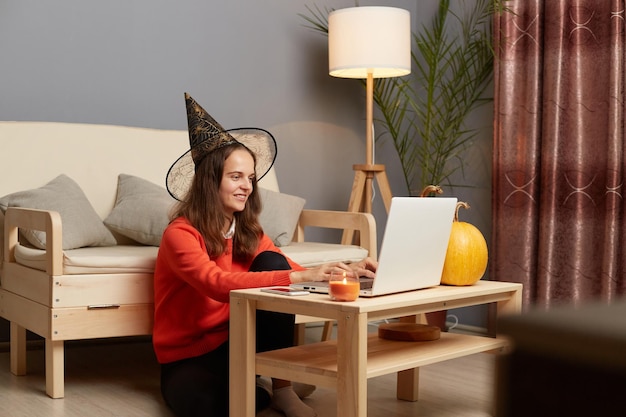 Indoor shot of smiling happy woman wearing witch hat combines celebrating halloween and working online on laptop sitting on laptop and typing on keyboard