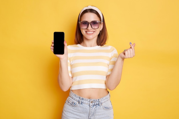 Indoor shot of smiling lovely young woman wearing striped tshirt posing isolated over yellow background showing cell phone with empty display and korean love gesture