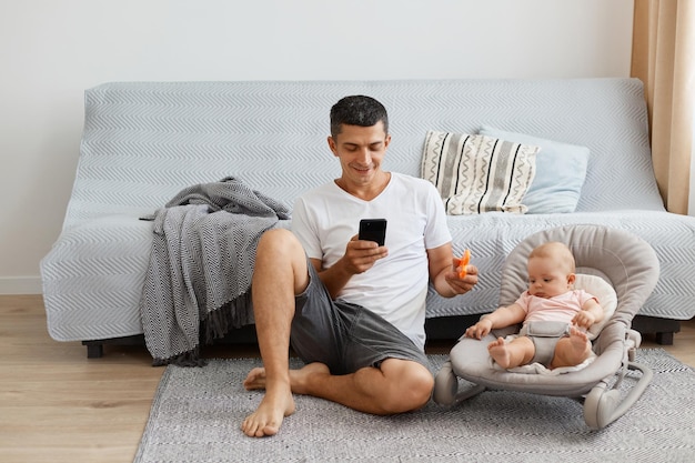 Indoor shot of smiling man wearing white casual style Tshirt and jeans short sitting on floor with daughter in rocking chair father using smart phone and showing toy for kid