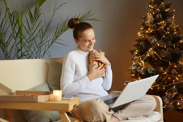 Indoor shot of smiling petty positive female with bun hairstyle sitting on sofa near Christmas tree and having video call via laptop and embracing present box celebrating New year Eve