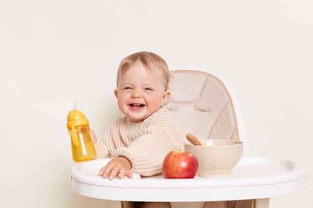 Indoor shot of smiling positive baby sitting in a chair against white isolated background kid with water bottle and fruit having fun during breakfast or dinner