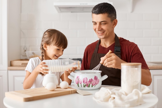 Indoor shot of smiling positive father sitting at table with daughter and cooking together in kitchen little girl pouring milk into pot and smiling will making pancakes