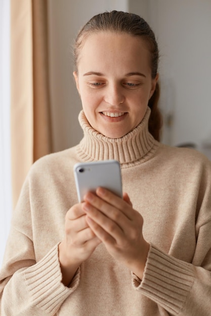 Indoor shot of smiling positive woman with ponytail hairstyle wearing beige casual style sweater, standing with mobile phone in hands, checking social networks, writing message.