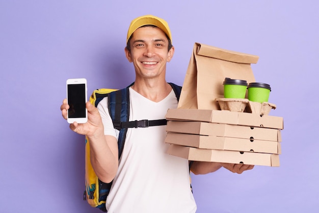 Indoor shot of smiling satisfied man showing blank screen of mobile phone and holding pizza boxes coffee to go presenting area for advertisement isolated on purple background