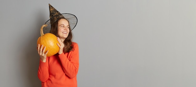 Indoor shot of smiling satisfied woman holding big ripe orange pumpkin wearing sweater and witch hat looking away at copy space for advertisement isolated over gray background