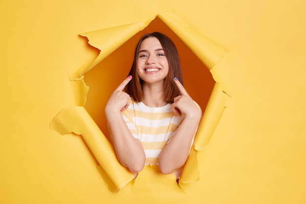 Indoor shot of smiling young adult woman stands in torn paper hole pointing at her perfect smile and teeth looking through breakthrough of yellow background