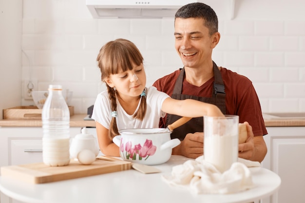 Indoor shot of smiling young dark haired man wearing maroon t shirt and apron cooking in kitchen with little daughter, preparing dough for pie or cake for birthday,