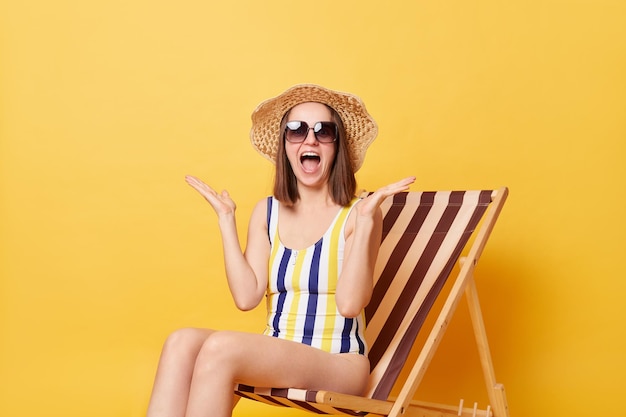 Indoor shot of surprised amazed excited woman with slim body wearing straw hat sunglasses and swimsuit posing isolated over yellow background screaming with happiness rejoices her tour