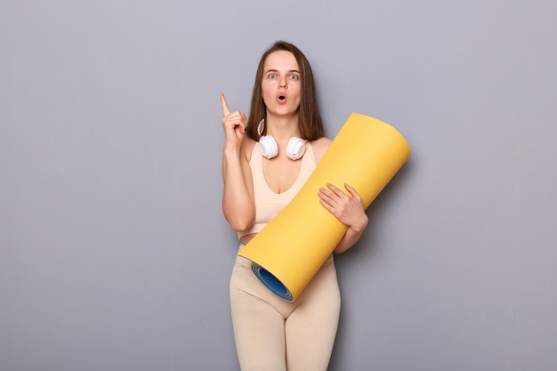 Indoor shot of surprised woman trainer wearing sportswear standing with karemat in hands and raising finger having idea of new set of workout isolated over gray background