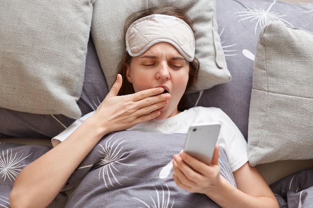 Indoor shot of tired sleepy young woman wearing sleeping mask lying in comfortable bed on pillow and using cell phone after waking up yawning checking social network