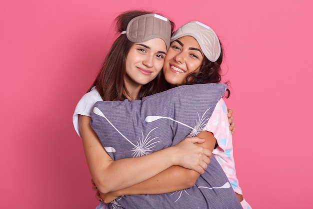 Indoor shot of two beautiful attractive happy girls, smiling sincerely while looking at camera and hug each other, wearing sleeping masks and pajamas, embracing pillow against rose wall.