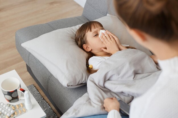 Indoor shot of unhealthy ill female child with handkerchief lying on sofa under blanket suffering runny nose and high temperature mother sitting near her little sick girl