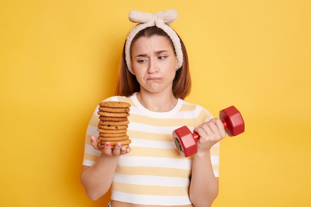 Indoor shot of upset pensive woman wearing t shirt and hair band posing isolated on yellow background holding dumbbell in one hand and looking at sweet cookie in another hand