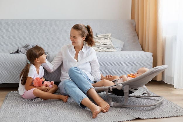 Indoor shot of woman wearing white shirt and jeans sitting on floor near sofa mother playing and communicating with her little children little girl with dark hair and infant baby in rocking chair