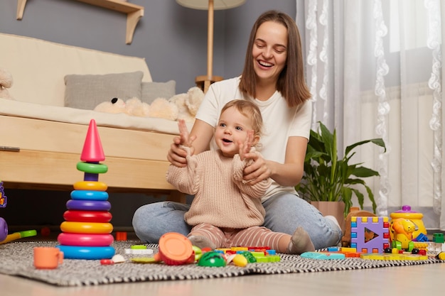 Indoor shot of woman with brown hair plays with baby in the room mother holding baby's hands teaching her to clap hands expressing happiness motherhood and childhood