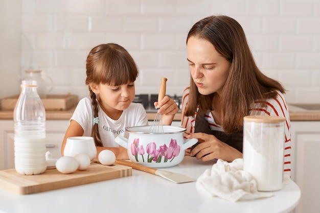 Indoor shot of woman with dark hair baking cookies or pastries with cute little preschooler girl in kitchen sitting at table and looking in pot with dough with frowning face disappointed with result