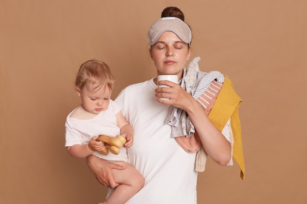 Indoor shot of young tired mom and little child after sleepless night exhausted woman with baby standing with coffee isolated over brown background Postpartum depression on maternity leave