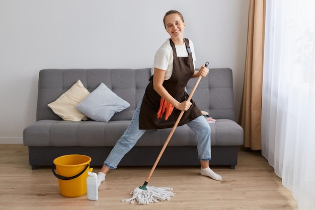 Indoor shot of young woman dancing while washing floor in living room holding mop as microphone having fun during house chores enjoying domestic duties cleaning her apartment