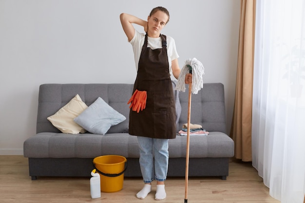 Indoor shot of young woman tired of cleaning house resting standing near sofa after washing floor with mop feeling pain in neck having exhausted expression